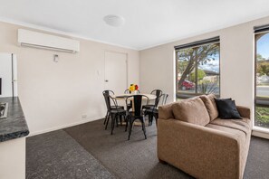 A modern living room with a beige sofa, a dining area with a table and chairs, and a large window with a view of the outside. - Arkana Motor Inn & Terrace Apartments (Mount Gambier)