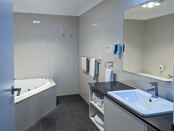 A modern bathroom with a bathtub, a sink, and a large mirror. There are towels hanging on hooks and a soap dispenser on the wall.