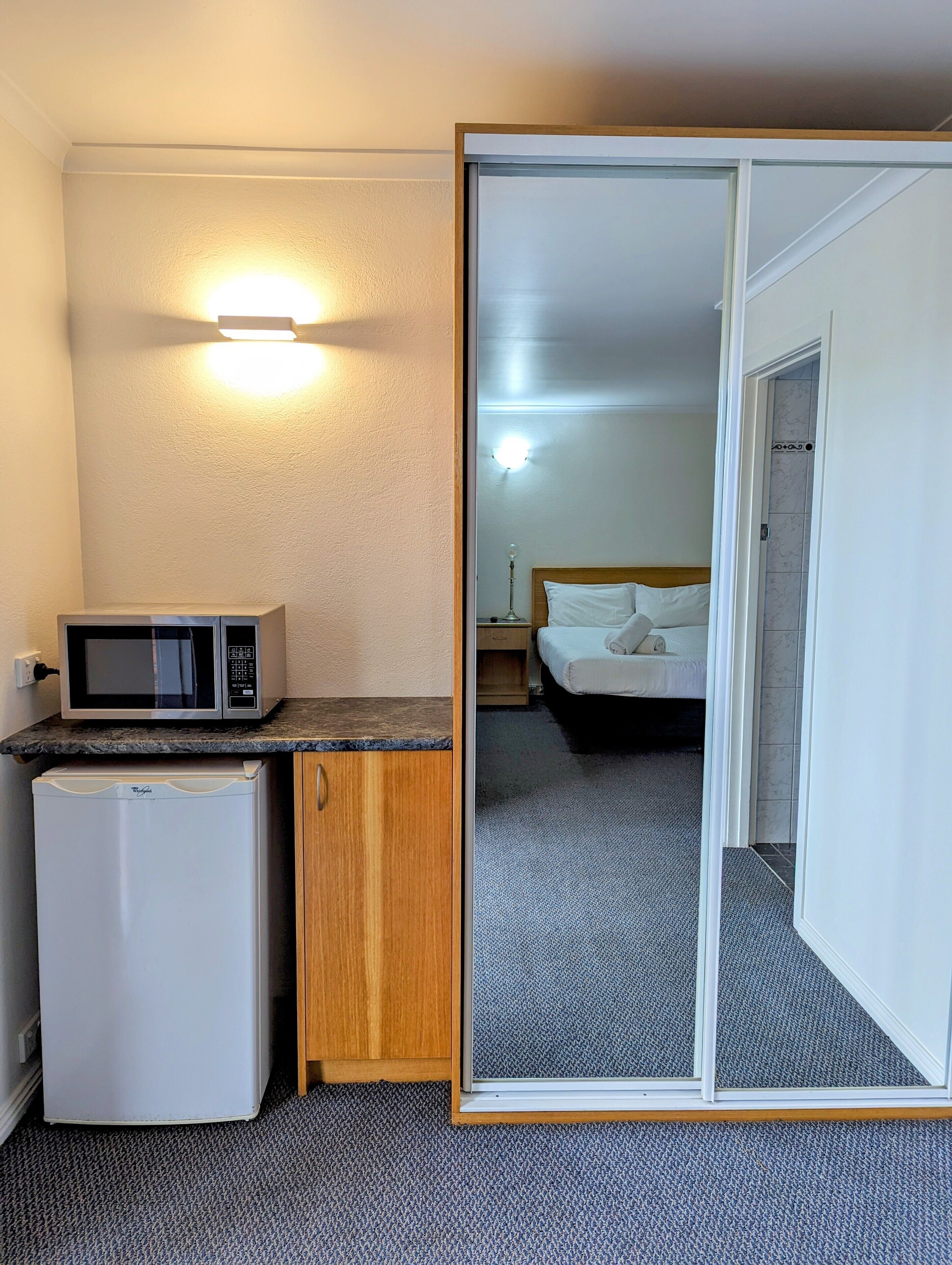 A compact hotel room with a kitchenette, a bed, and a bathroom visible through a sliding door.
