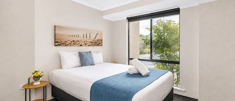 A hotel room with a large bed, a small side table with a vase of flowers, a painting on the wall, and a window with a view of greenery.