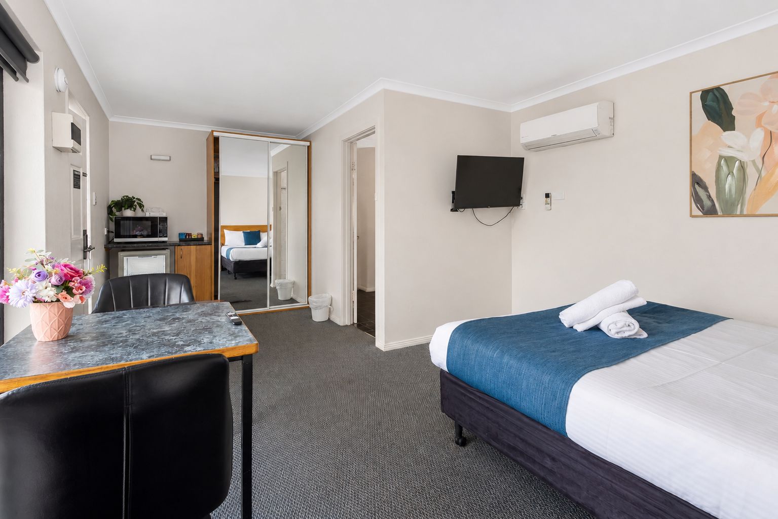 Habitación de hotel con cama, escritorio, silla, televisor montado en la pared y una pequeña cocina en el fondo.