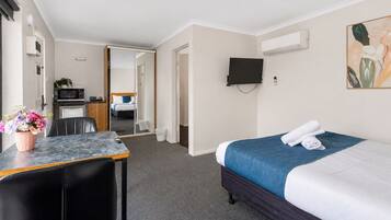 Habitación de hotel con cama, escritorio, silla, televisor montado en la pared y una pequeña cocina en el fondo.