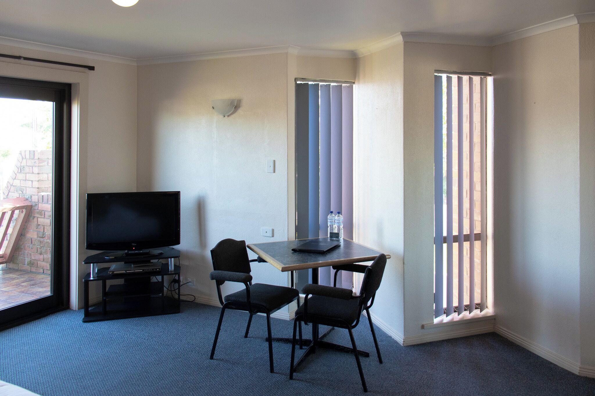 A small room with a TV on a stand, two chairs, a table, and a window with blinds.