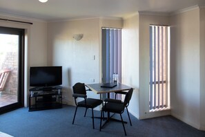 Une petite pièce avec un téléviseur posé sur un meuble, deux chaises, une table et une fenêtre avec des stores.