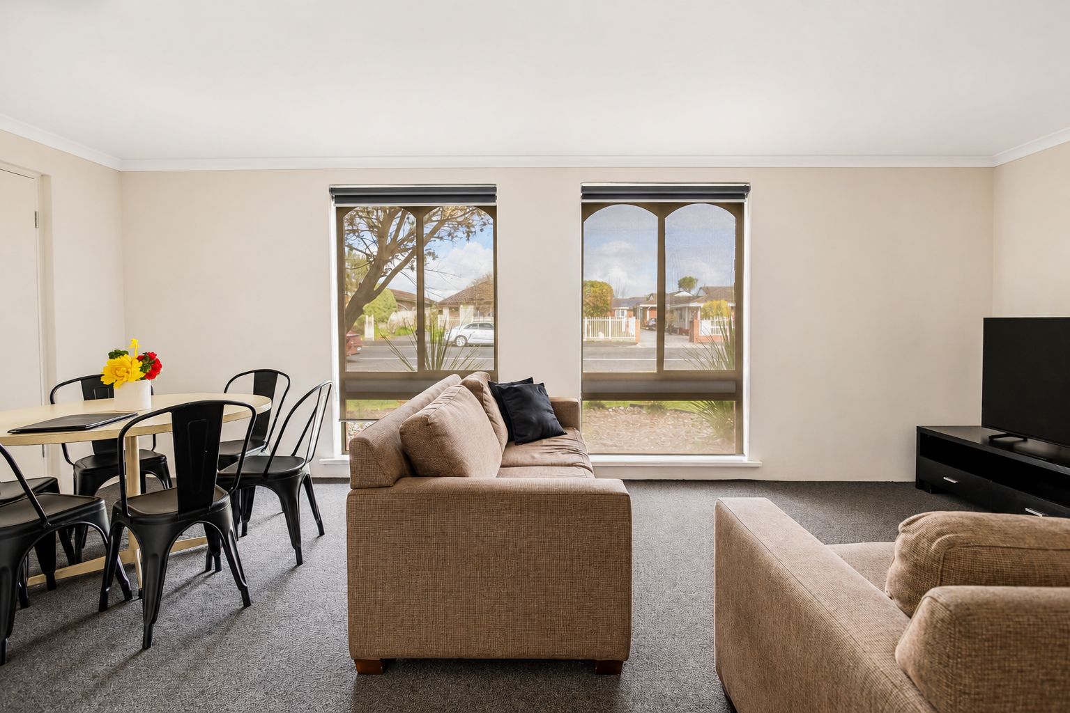 A modern living room with a sofa, a dining table with chairs, a television, and large windows with a view of a residential area.
