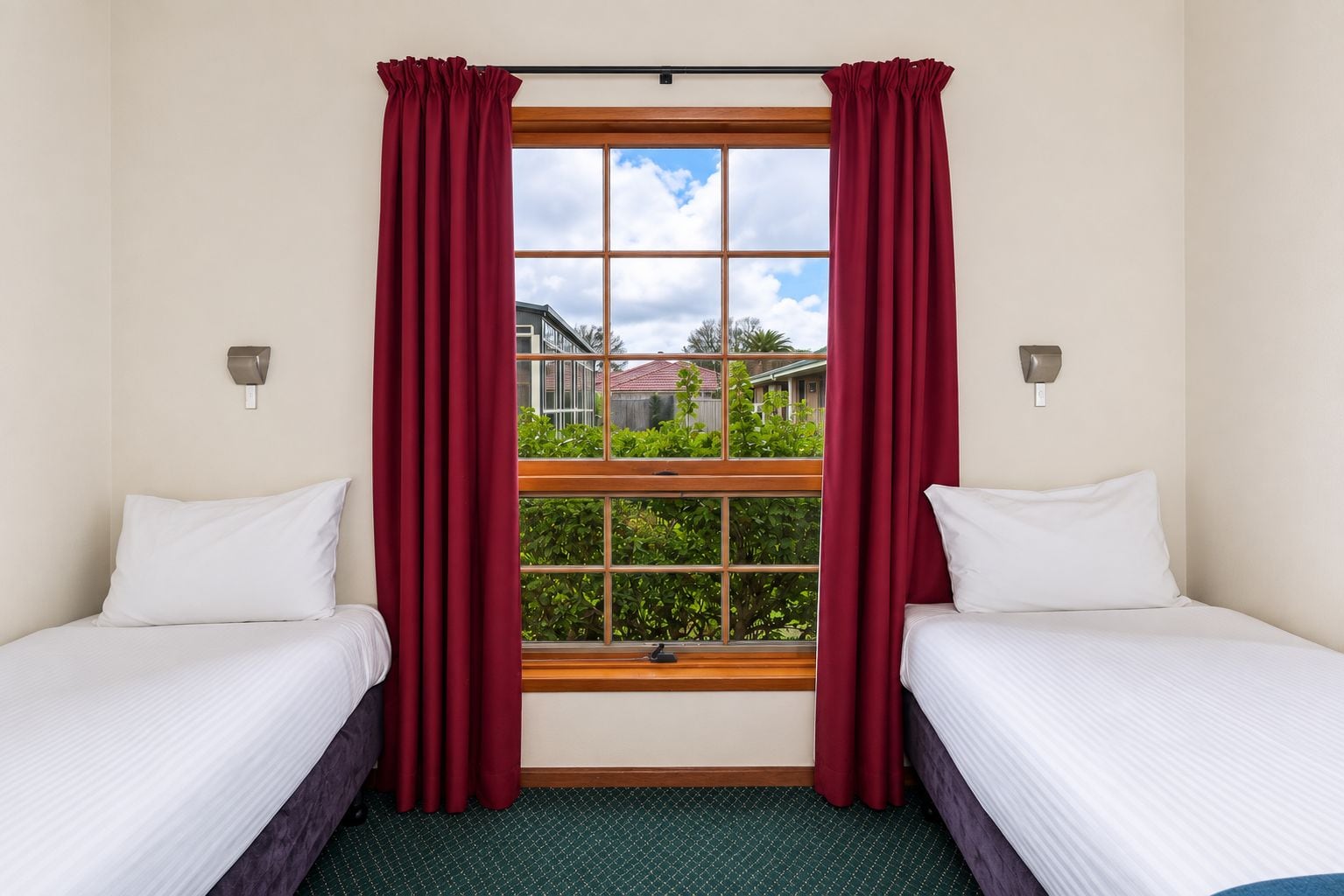 A hotel room with two beds, a large window with curtains, and a view of a residential area.