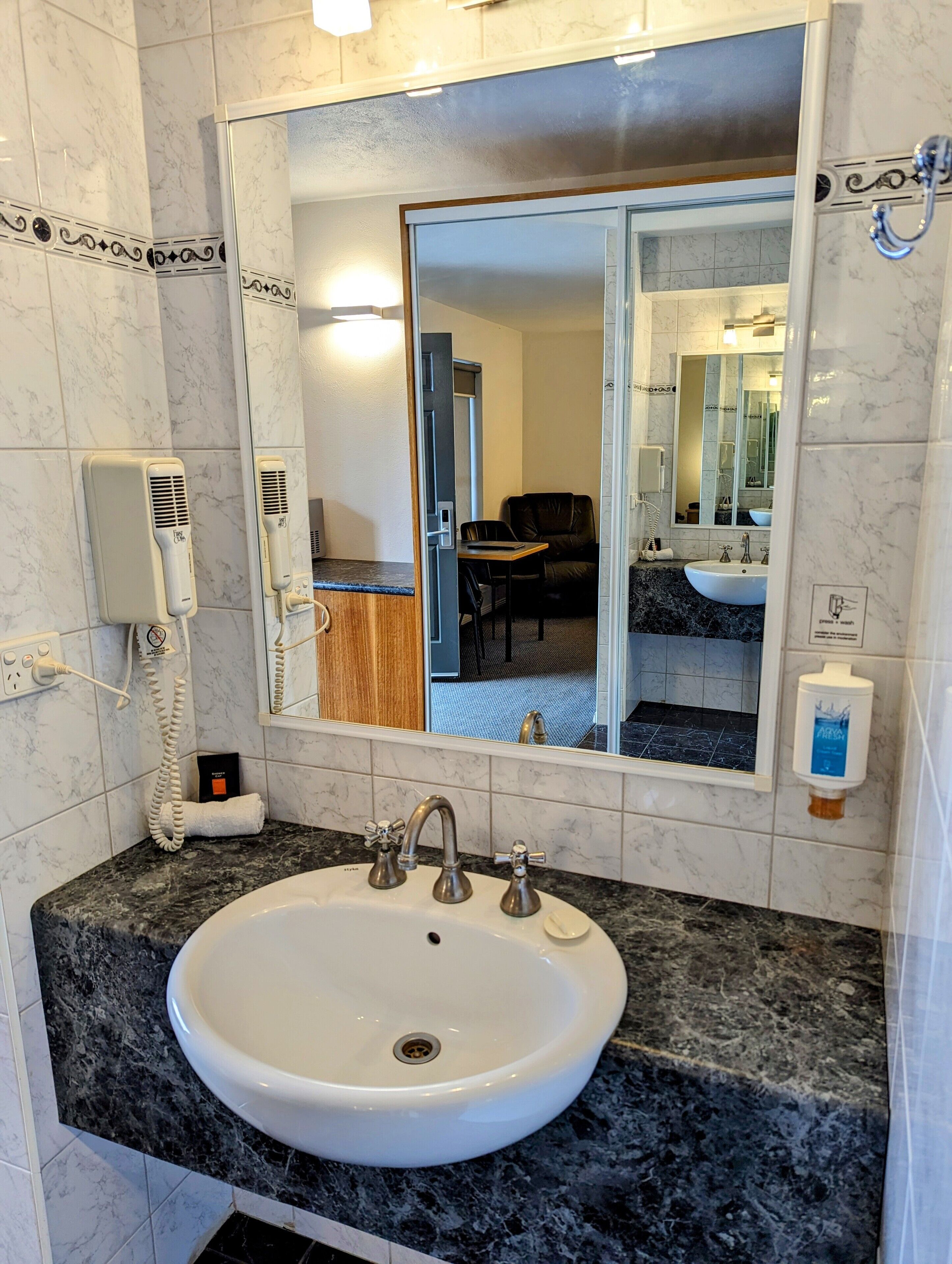 A bathroom with a white sink, marble tiles, a mirror, a hairdryer, and a hand soap dispenser.