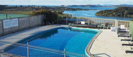 A pool area with a view of a lake and mountains, surrounded by a fence and lounge chairs.