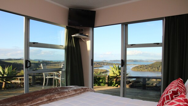 Un dormitorio con una cama grande, televisión y balcón con vistas a un lago y colinas.