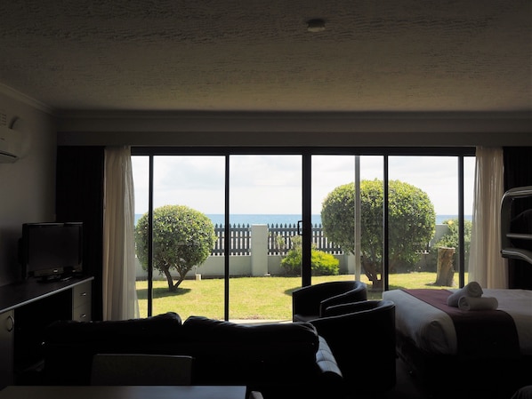 Quarto de hotel com uma grande porta de correr de vidro que dá para uma varanda com vista para o mar e um jardim cercado.