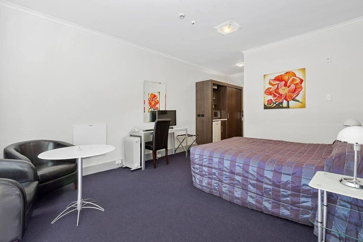 A hotel room with a bed, a desk with a chair, a small table, a radiator, and a painting on the wall.