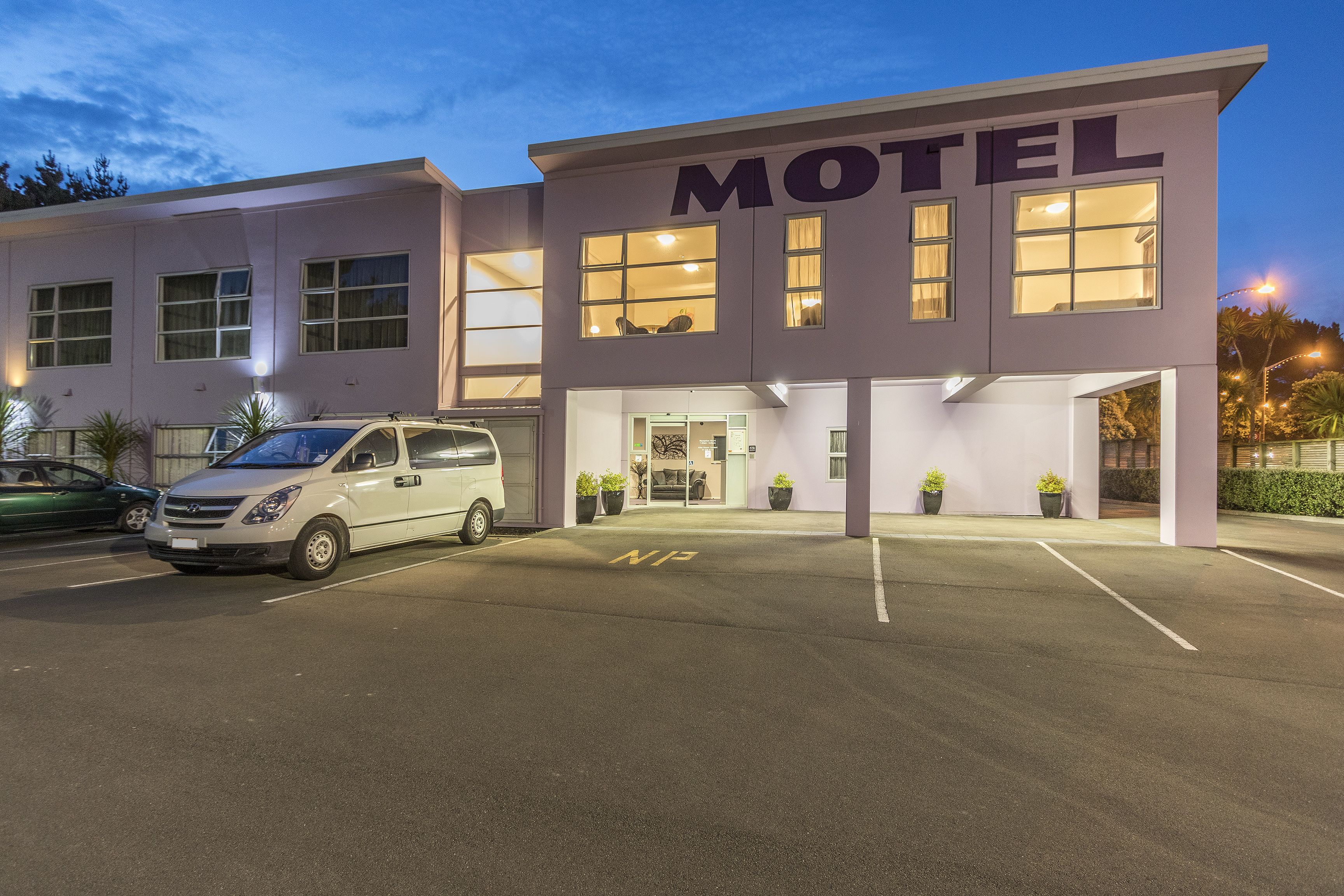 Een motel met een bord met de tekst MOTEL boven de ingang. Er staan potplanten voor het gebouw en een witte bestelwagen staat op de parkeerplaats.