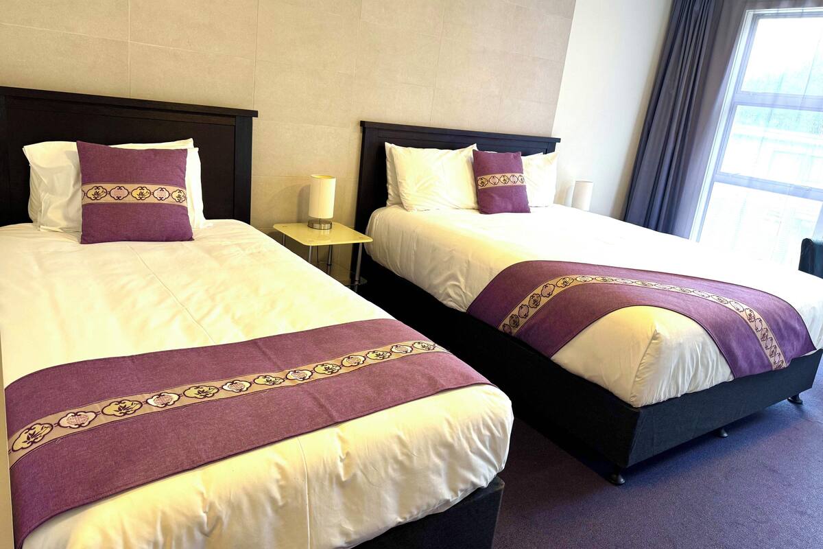 A hotel room with two beds, each with a headboard, a bedside table with a lamp, and a window with curtains.