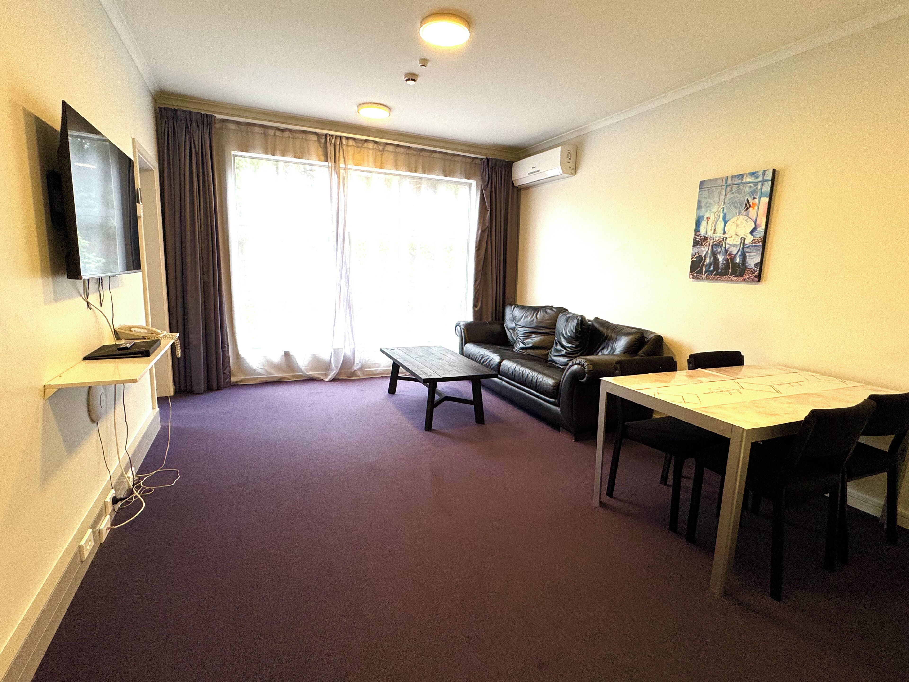 Una sala de estar con sofá, mesa de centro y mesa de comedor. Hay un televisor montado en la pared y un equipo de aire acondicionado encima.