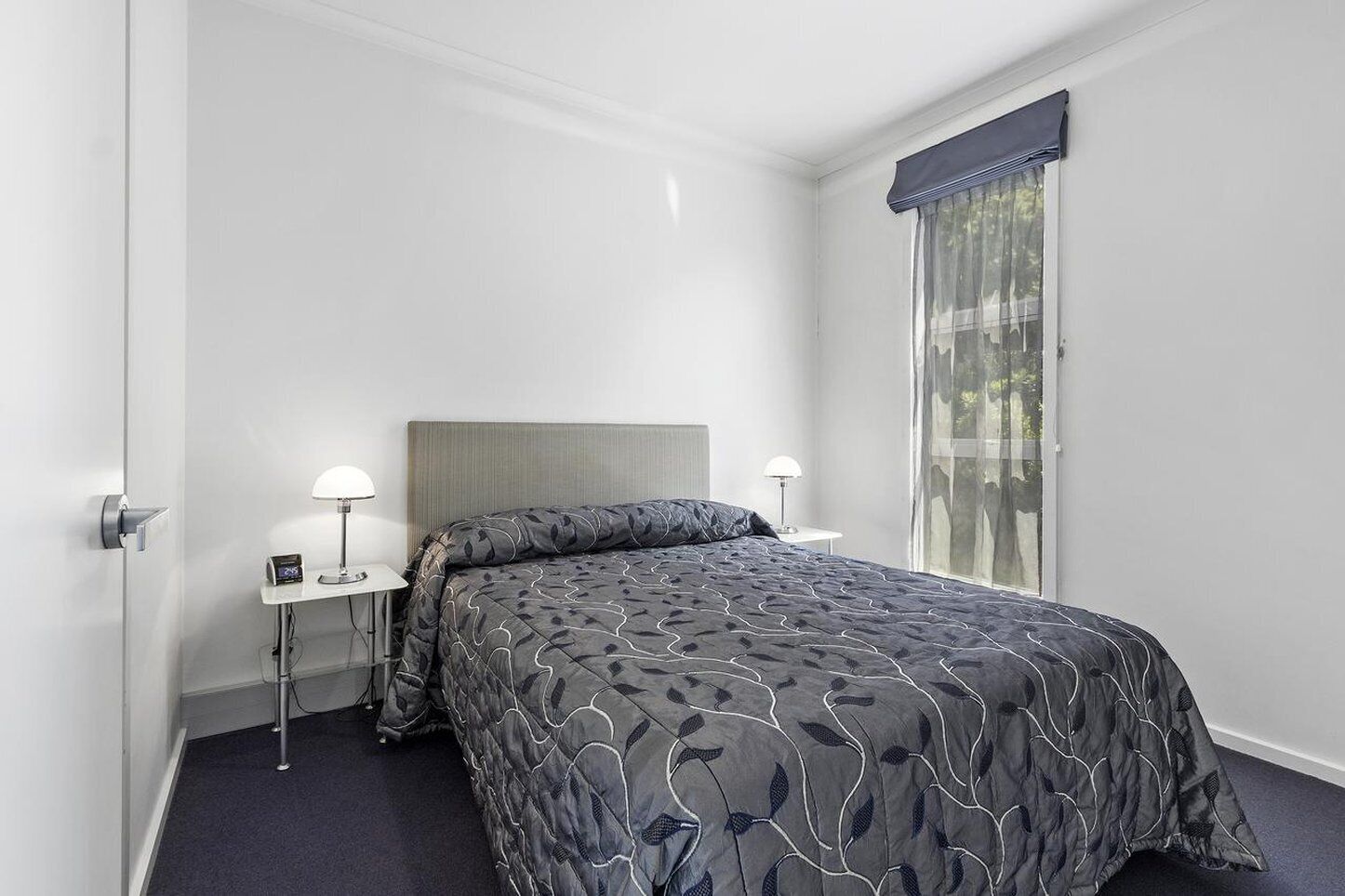 A modern hotel room with a large bed, two bedside tables with lamps, a window with a view, and a door.