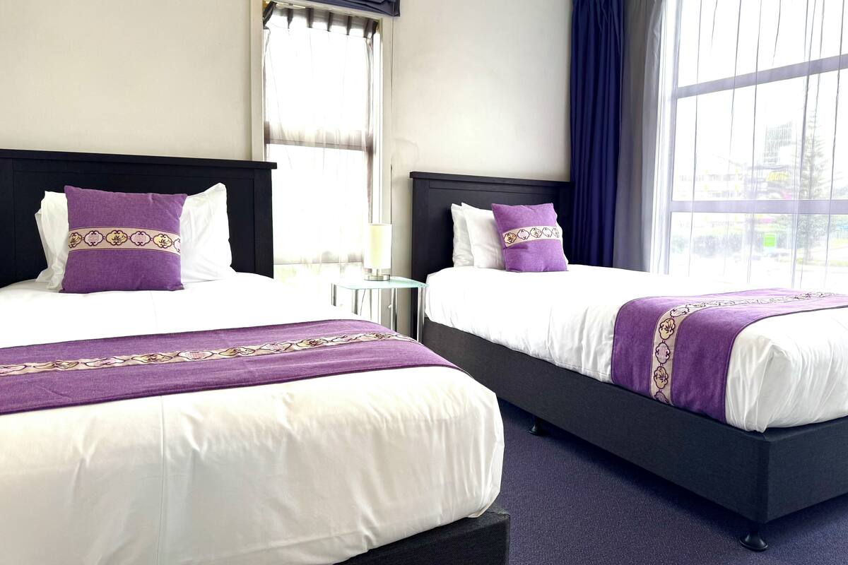 A hotel room with two beds, each with white linens and purple decorative pillows. There is a small bedside table with a lamp, and a large window with blue curtains.