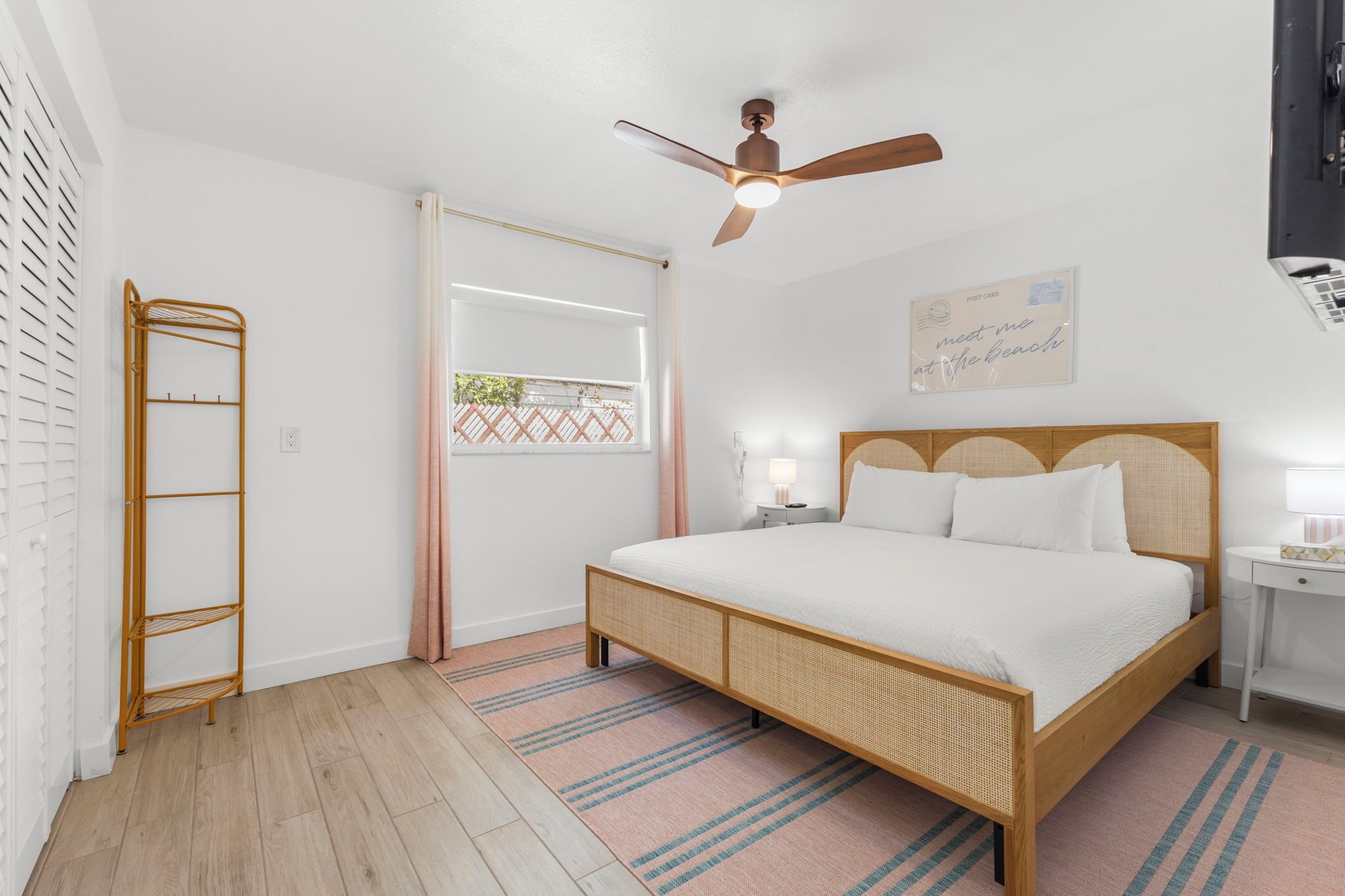 Un dormitorio con cama, ventilador de techo, mesita de noche, escritorio, ventana con cortinas y una postal colgada en la pared.