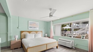 A bedroom with a bed, nightstands, a ceiling fan, a window with a view of outdoor furniture, and a wall-mounted air conditioner.