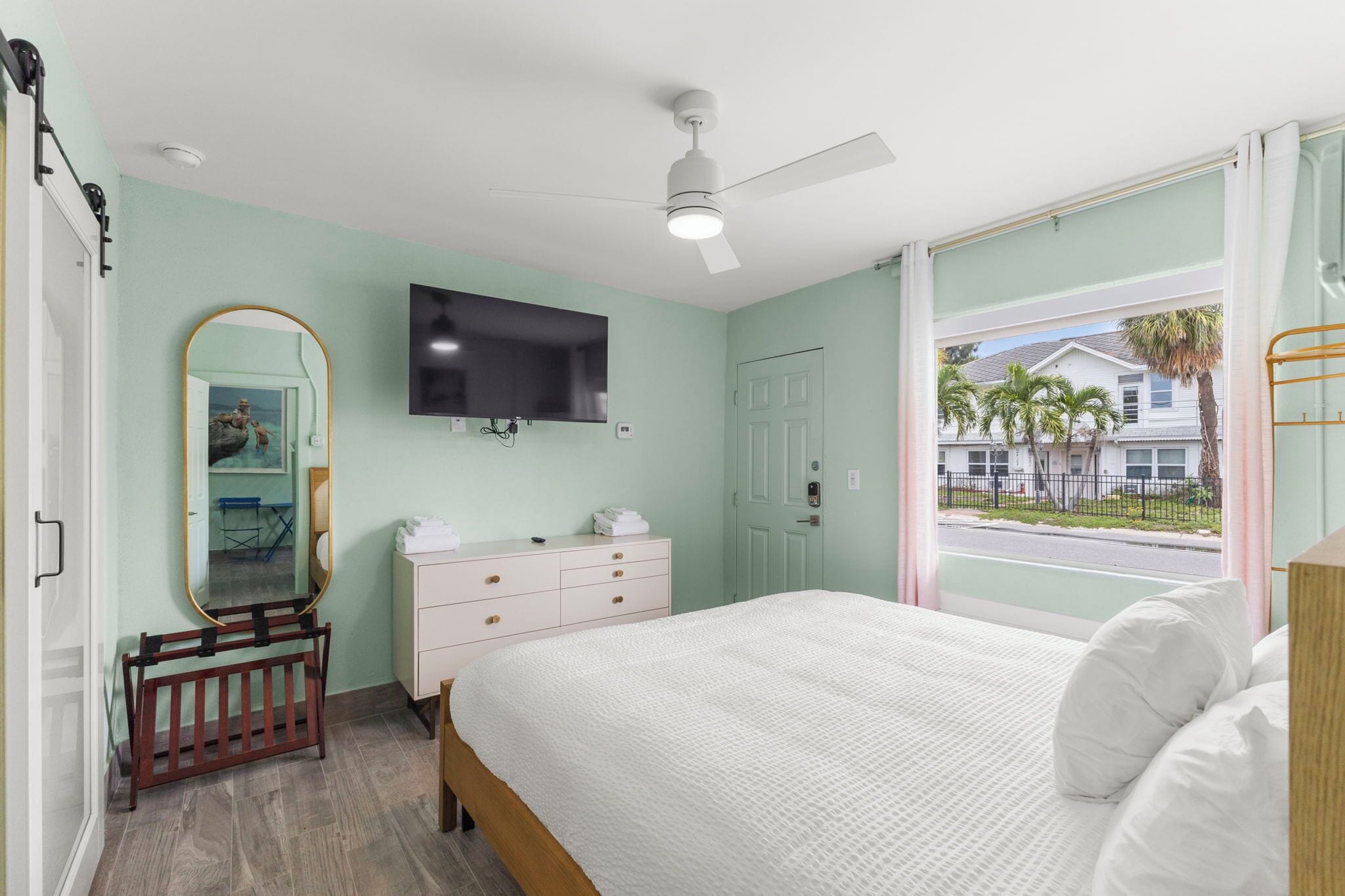 A bedroom with a bed, a ceiling fan, a TV, a window with a view of palm trees and a house, and a wooden crib.