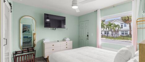 A bedroom with a bed, a ceiling fan, a TV, a window with a view of palm trees and a house, and a wooden crib.