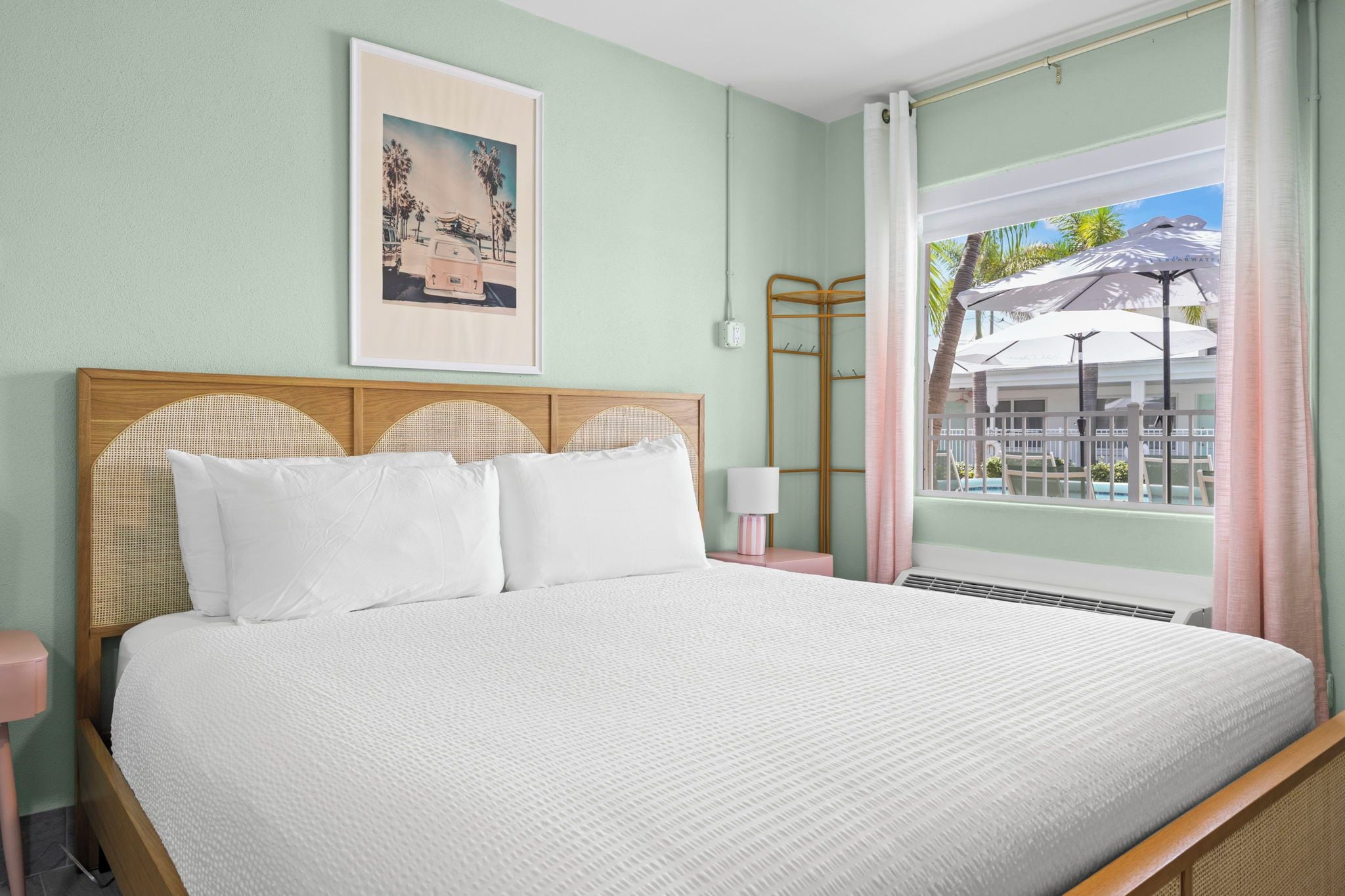 Ein Bett mit weißer Bettwäsche, einem hölzernen Kopfteil, einem Wandbild und einem Fenster mit Blick auf Palmen und ein Gebäude.