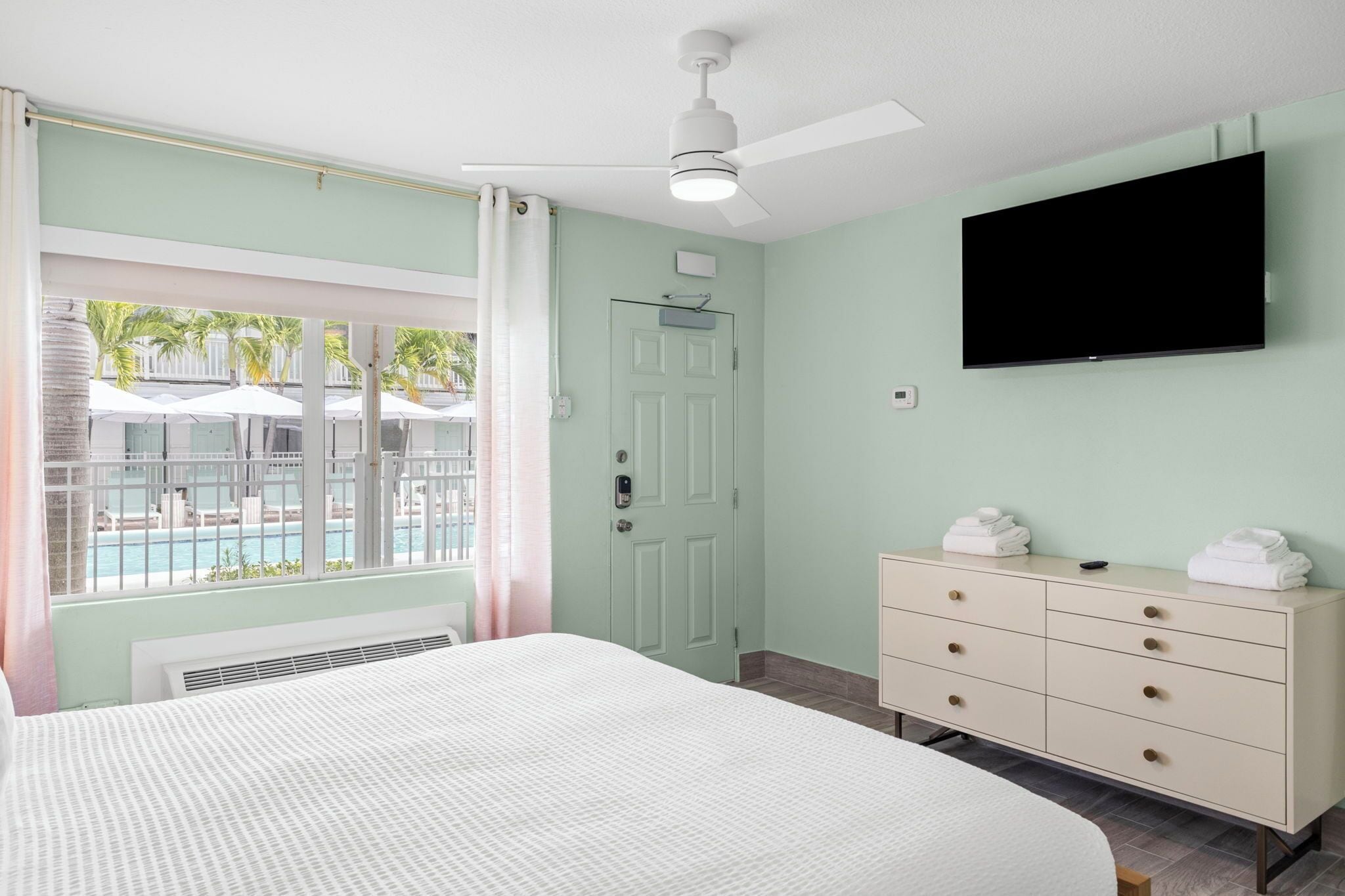 A hotel room with a bed, a dresser, a TV, a ceiling fan, a window with a view of a pool, and a door.