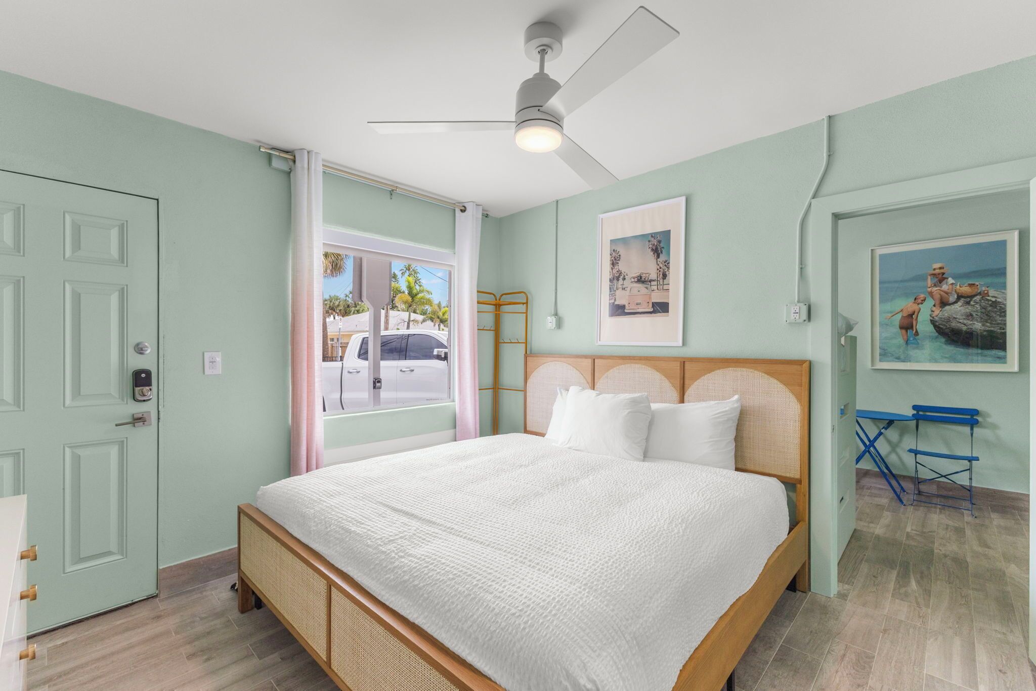 A bedroom with a bed, a ceiling fan, a window with a view of the street, and a door.