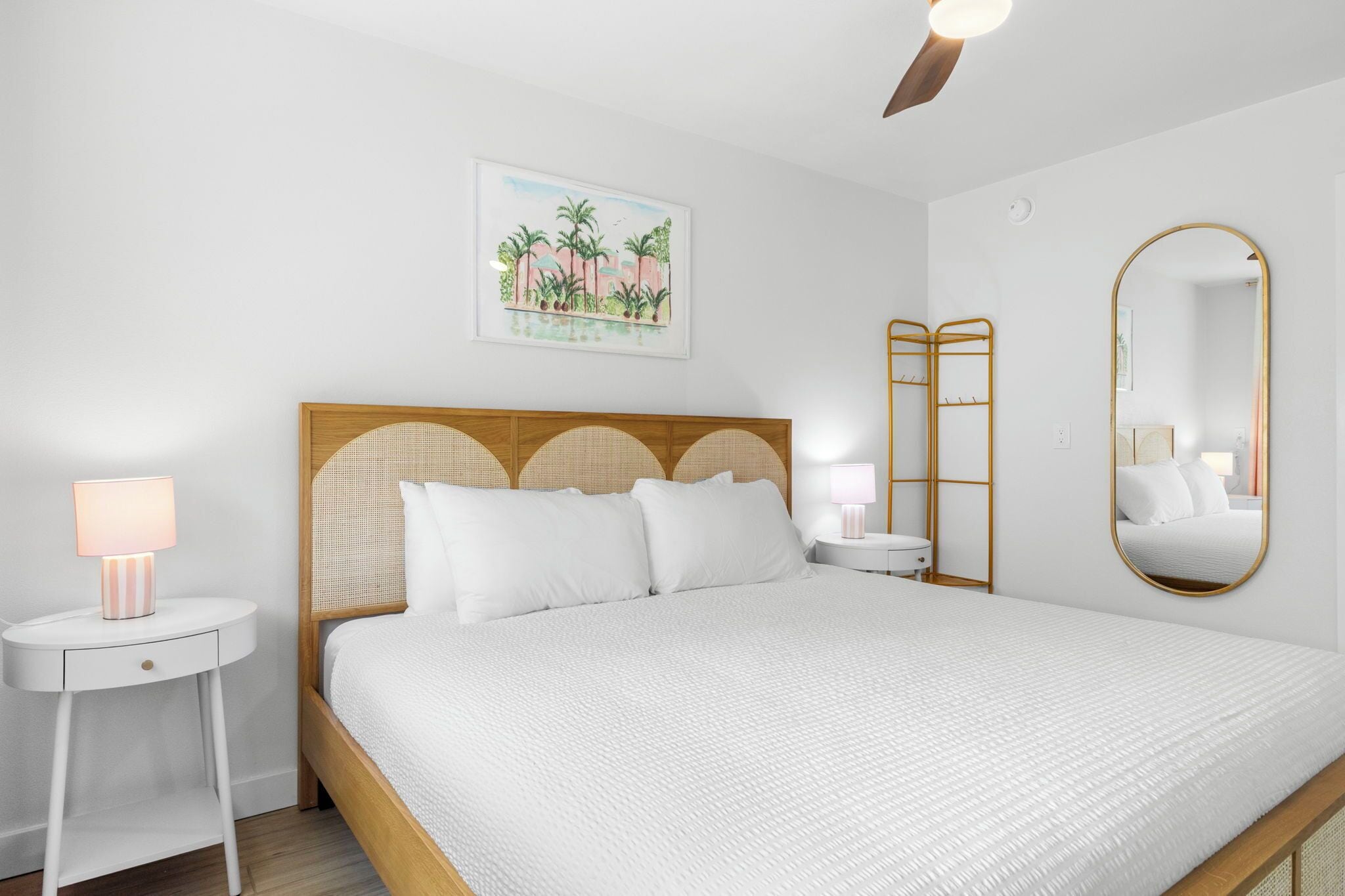 A neatly made bed with a wooden headboard, two bedside tables with lamps, a wall-mounted mirror, and a painting above the bed.