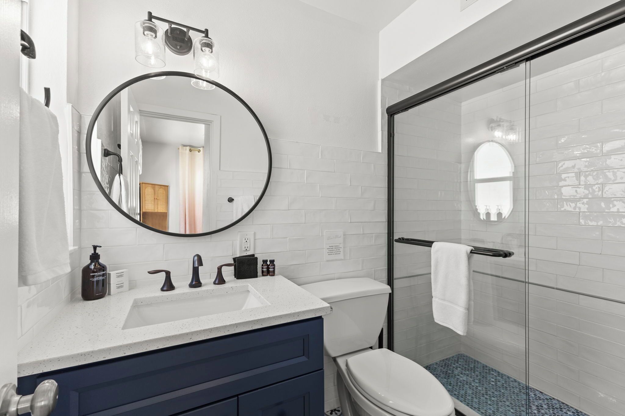 Ein modernes Badezimmer mit weiß gefliester Wand, einem runden Spiegel, einem Waschbecken mit zwei Wasserhähnen, einer Toilette und einer Duschkabine aus Glas.