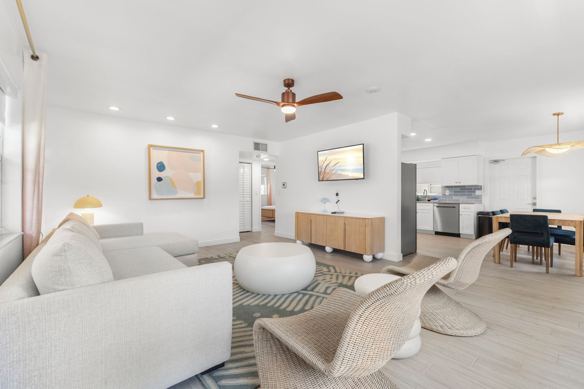 A modern living room with a sofa, ottoman, and wicker chairs. There is a dining area with a table and chairs, and a kitchen in the background.