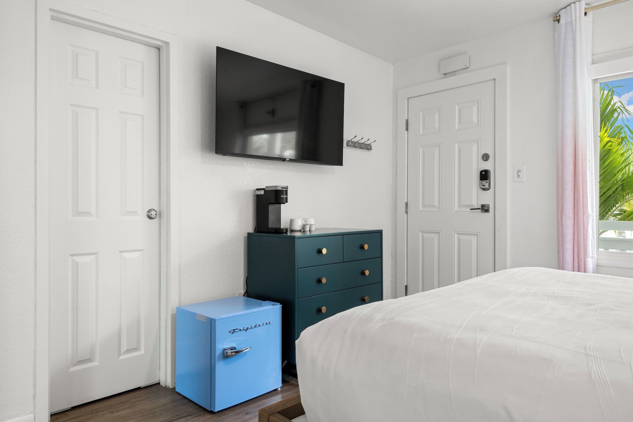 A hotel room with a bed, a TV mounted on the wall, a coffee machine, a blue storage unit, and a door with a lock.