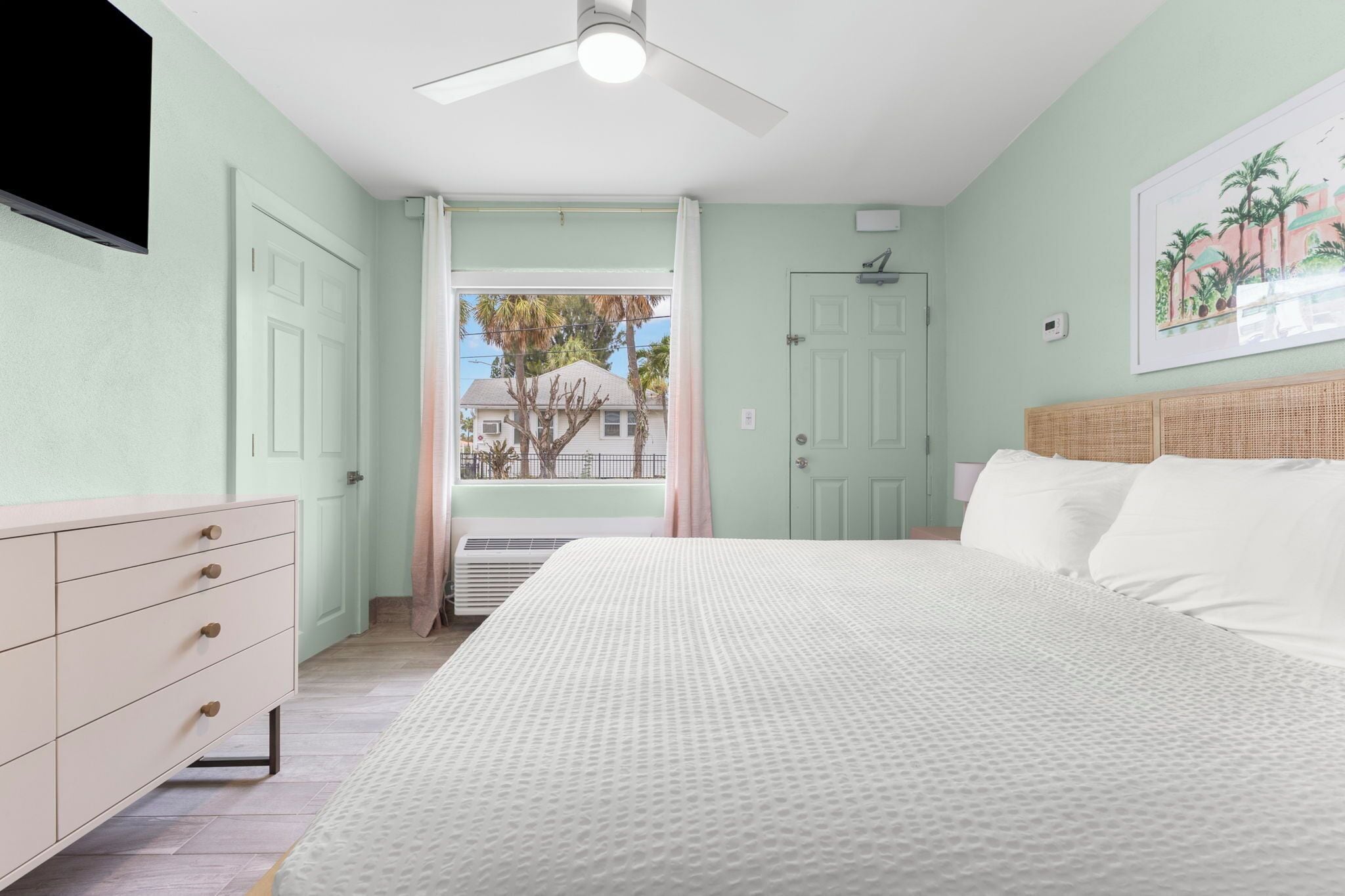 A bedroom with a bed, dresser, ceiling fan, and a window with a view of a house and palm trees.