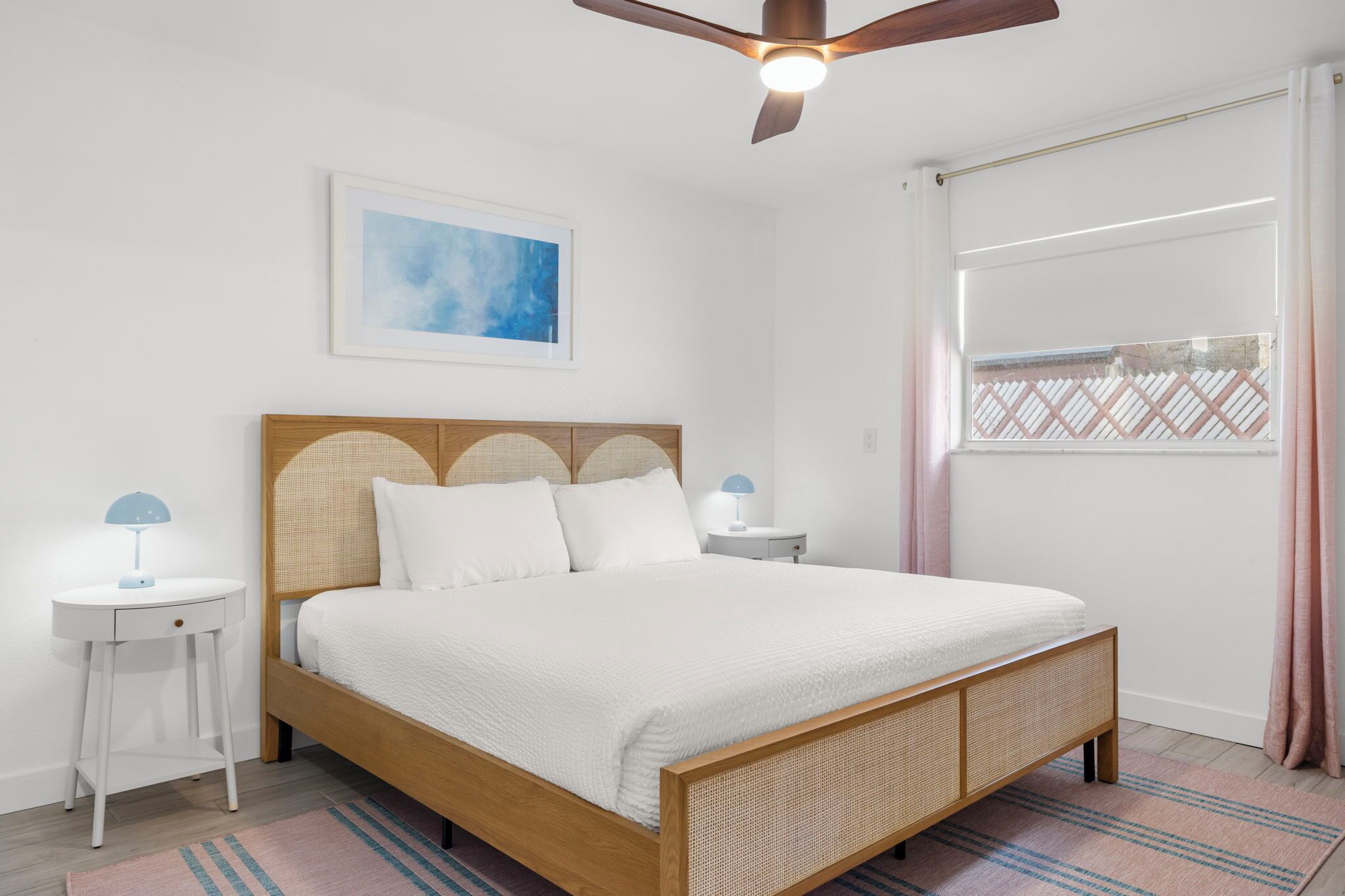 Un dormitorio con cama, ventilador de techo, mesita de noche, lámpara, un cuadro enmarcado y una ventana con cortinas.