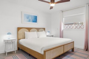 Un dormitorio con cama, ventilador de techo, mesita de noche, lámpara, un cuadro enmarcado y una ventana con cortinas.