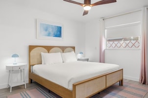 Un dormitorio con cama, ventilador de techo, mesita de noche, lámpara, un cuadro enmarcado y una ventana con cortinas.