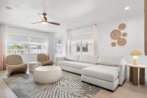 Uma sala de estar moderna com um sofá, cadeiras, um banquinho redondo, um ventilador de teto e uma vista através de grandes janelas.