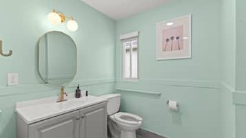 A bathroom with a white sink, gold faucet, and a toilet. There is a mirror with lights above it, a window with a view of palm trees, and a framed picture on the wall.