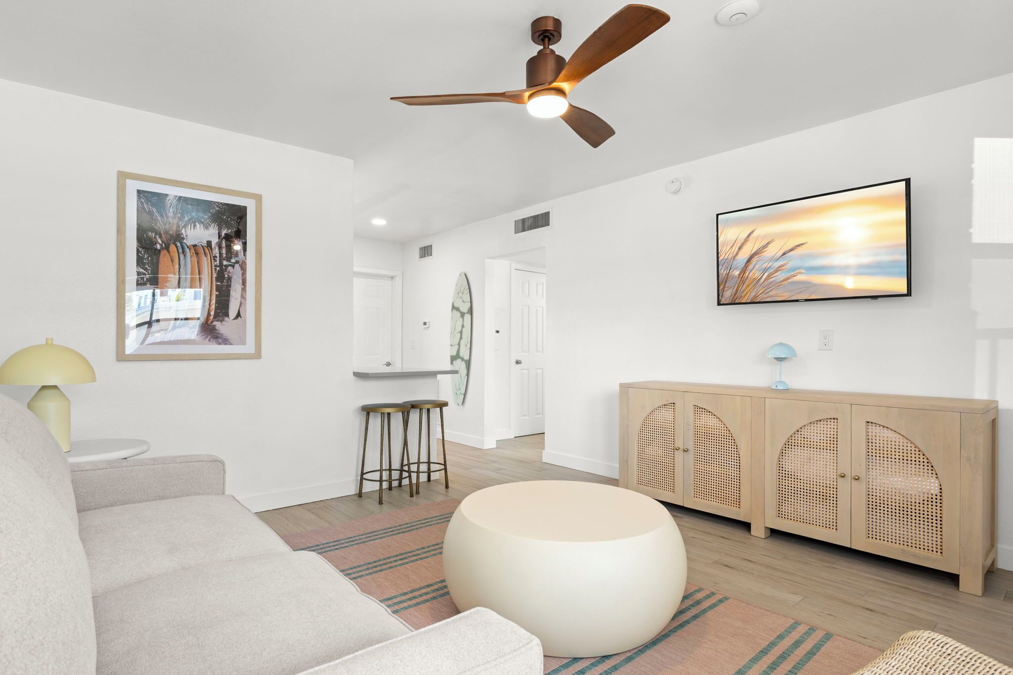 Un salon moderne avec un canapé, un pouf rond, un coin repas avec une table et des chaises, une cuisine avec une porte blanche et un ventilateur de plafond.