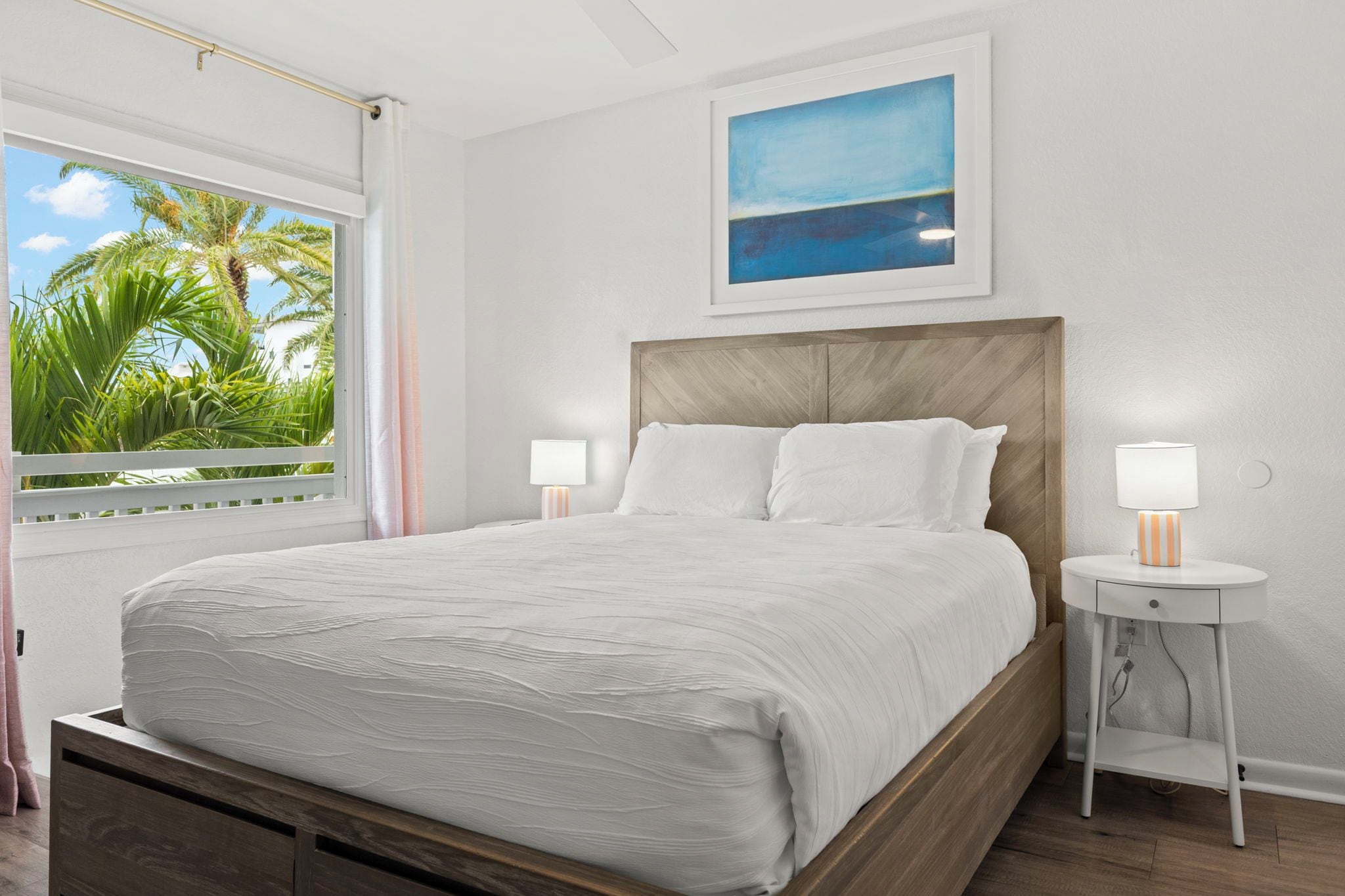 A bedroom with a bed, two bedside tables, a window with a view of palm trees, and a painting on the wall.