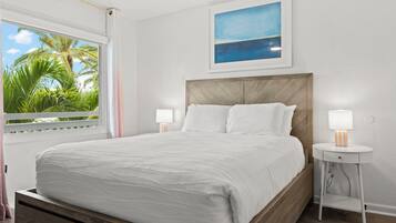 A bedroom with a bed, two bedside tables, a window with a view of palm trees, and a painting on the wall.