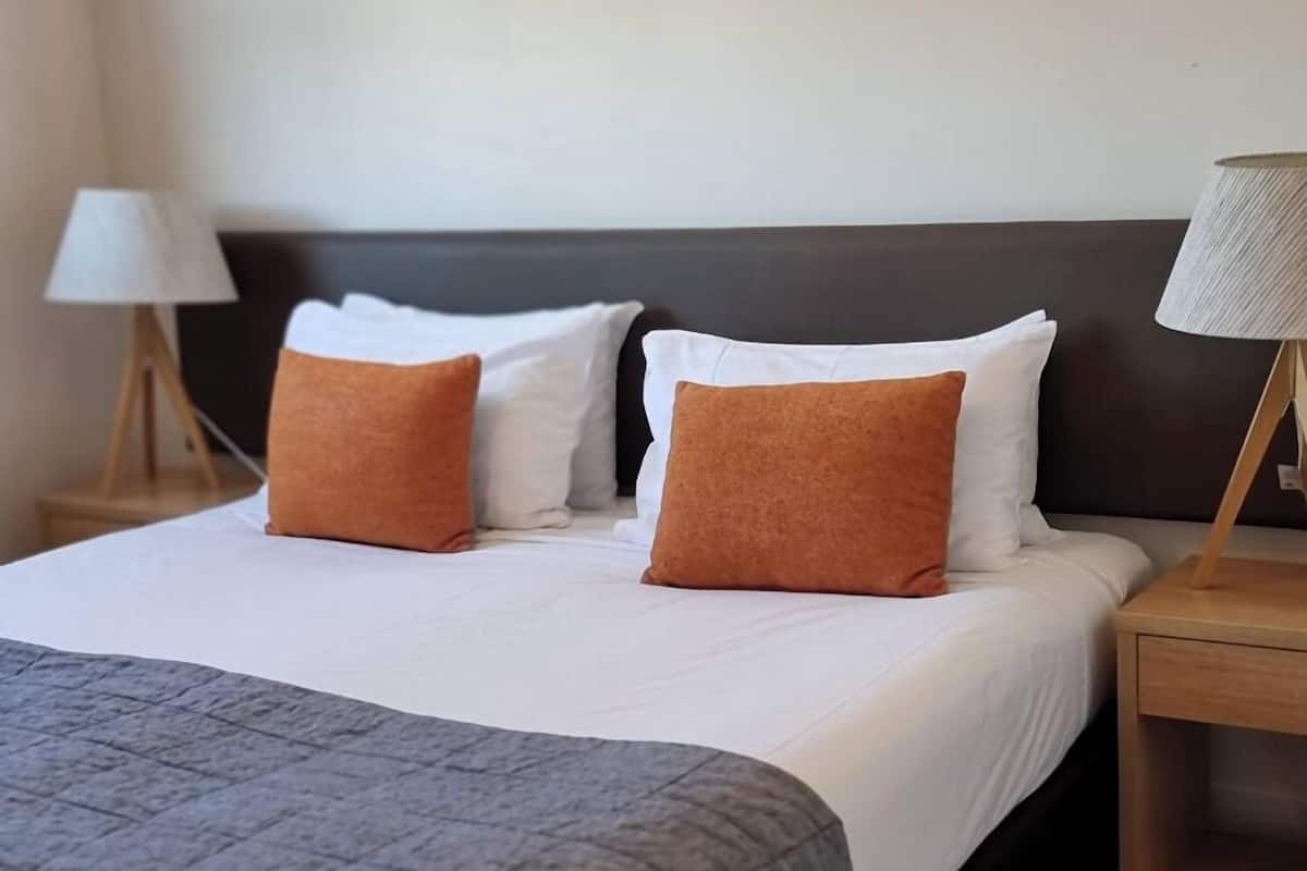 Una cama bien hecha con almohadas blancas y marrones, una manta gris y una mesita de noche de madera con una lámpara.