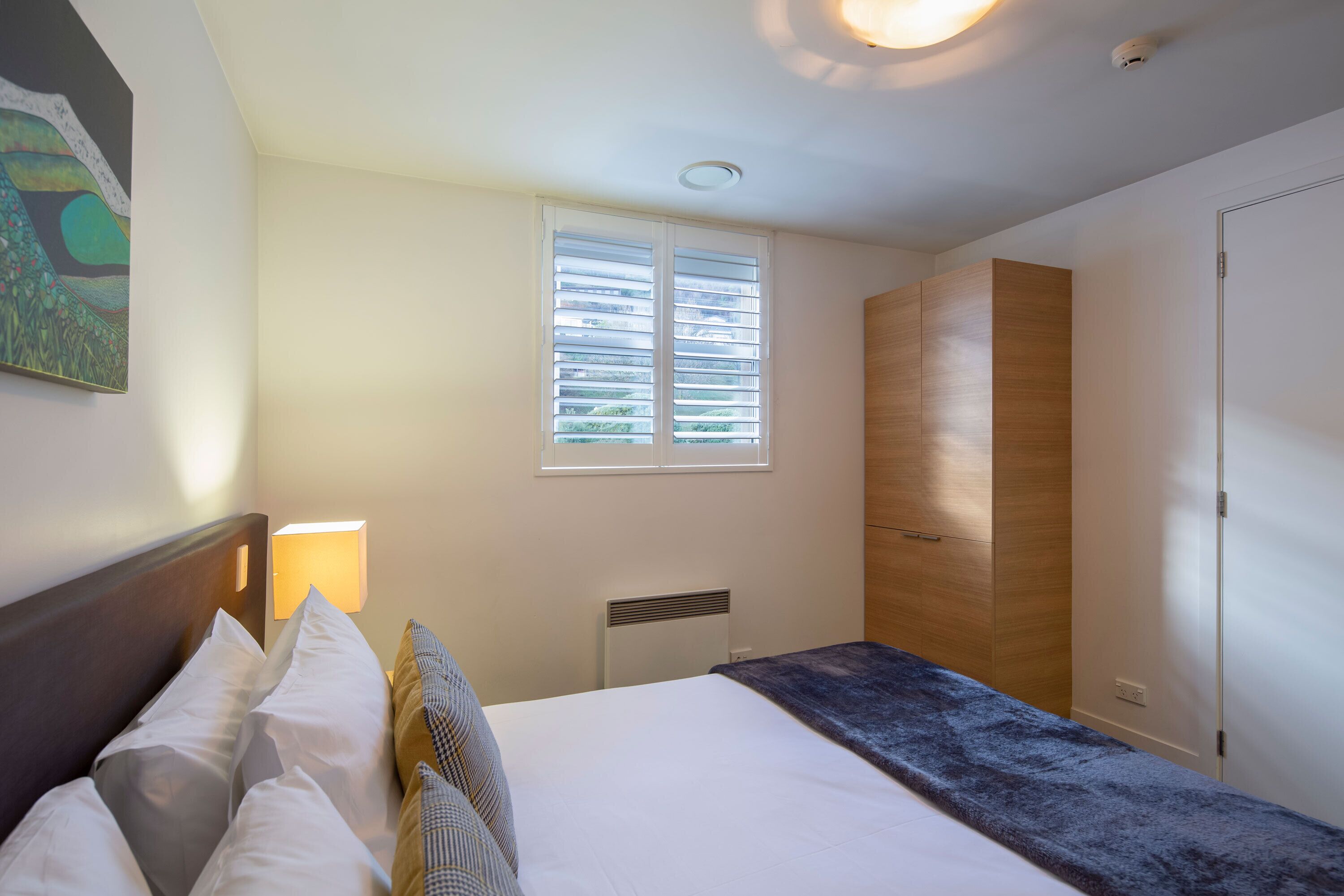 Une chambre à coucher comprenant un lit, une table de chevet, une fenêtre avec des stores, une armoire et un luminaire au plafond.