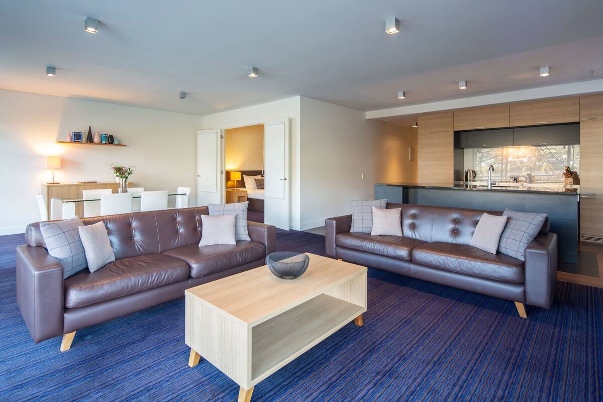 Una sala de estar moderna con dos sofás de cuero, una mesa de centro de madera y una zona de cocina con un ventanal grande.