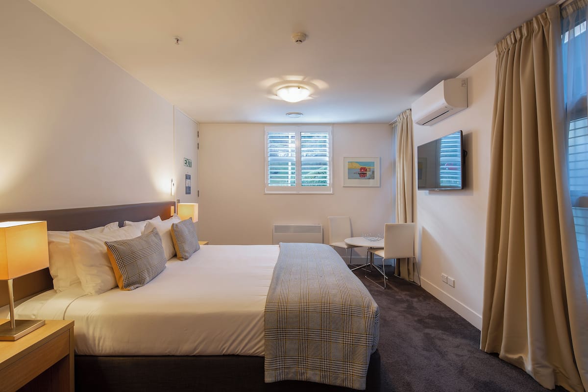 Habitación de hotel con cama, mesita de noche, lámpara, escritorio, silla, televisor, ventana y cortinas.