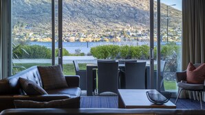 A room with a large window offering a view of a lake and mountains. Inside, there is a sofa, a table, and chairs. - Marina Apartments Element Escapes (Queenstown)