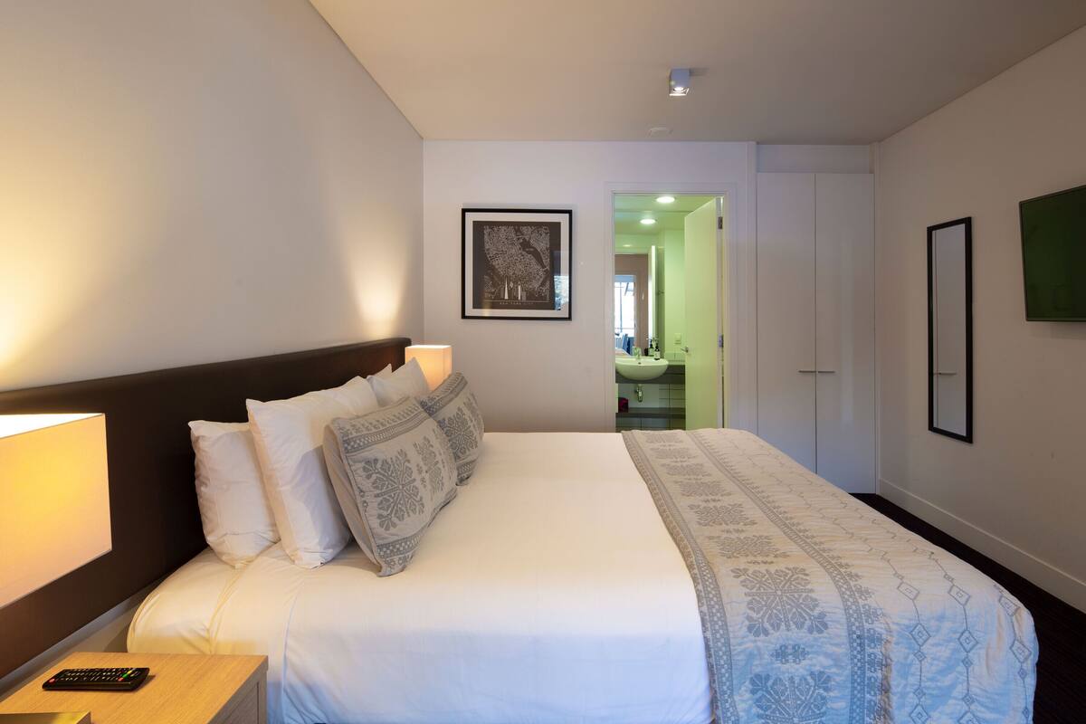 Una cama bien hecha con almohadones estampados, una lámpara de noche y un control remoto sobre una mesita de madera.