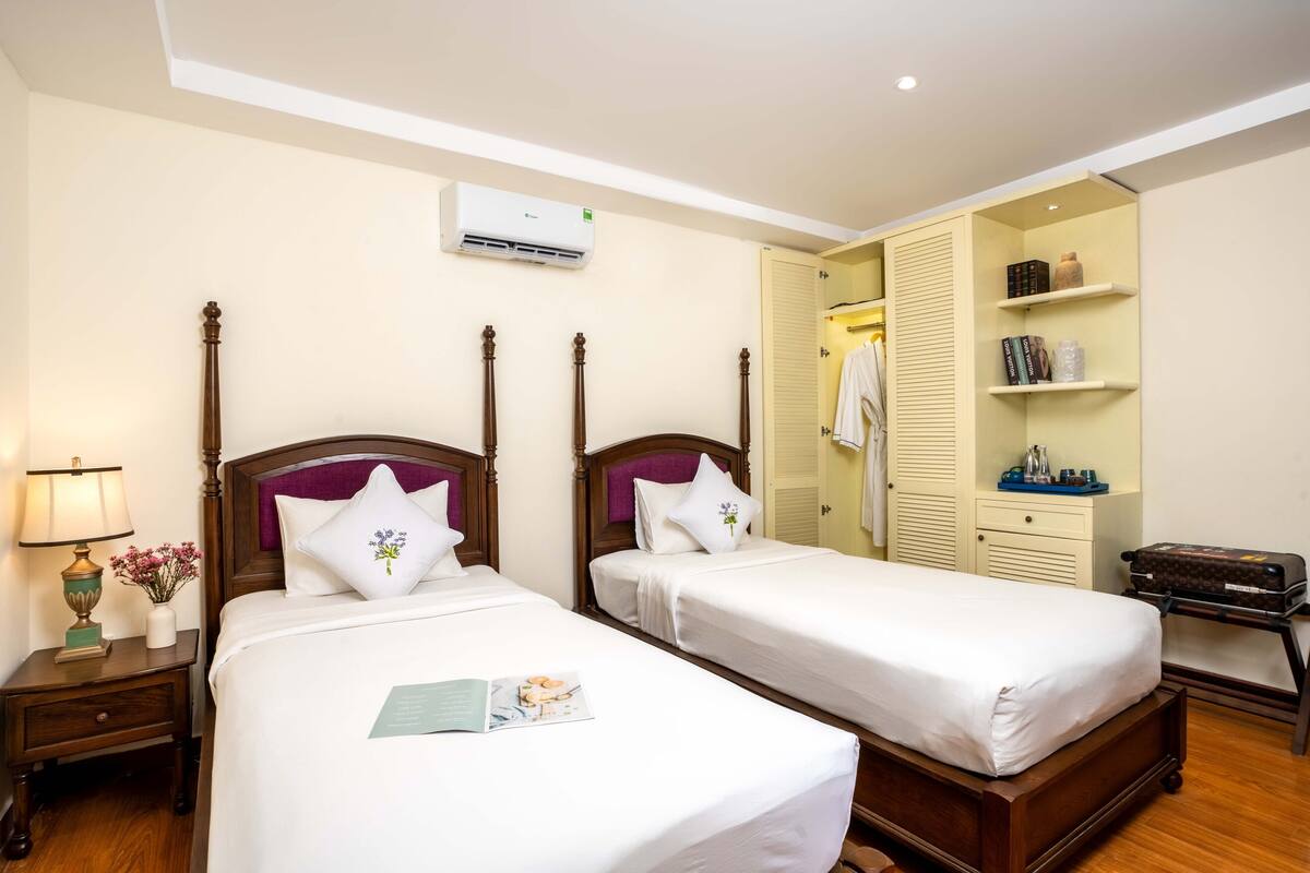 A hotel room with two single beds, a bedside table with a lamp, a small table with a vase of flowers, and a wardrobe with shelves.