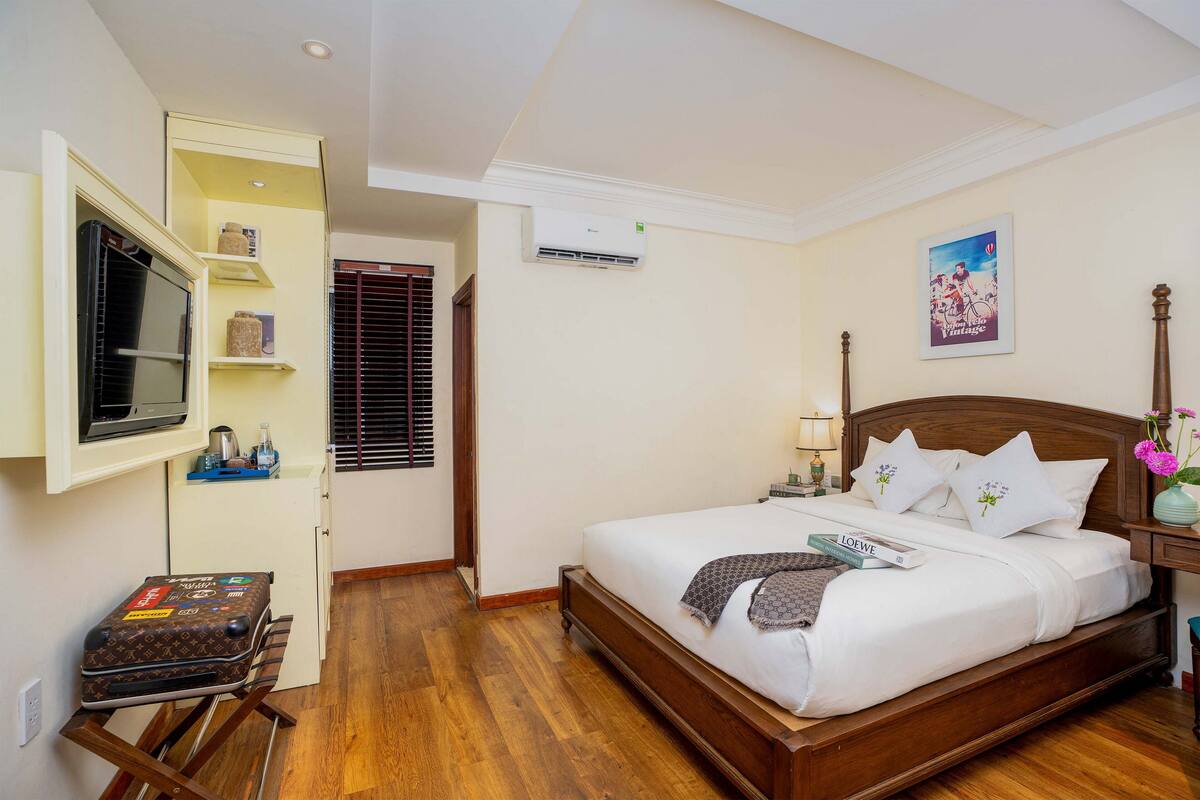 A neatly arranged hotel room with a wooden bed, a small desk with a suitcase, a television, and a window with blinds.