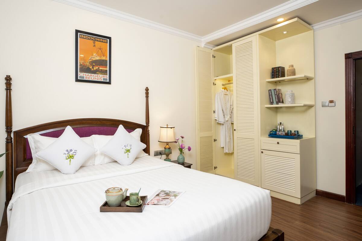 A neatly made bed with white bedding and decorative pillows, a bedside table with a lamp and a tray of items, and a wardrobe with shelves.