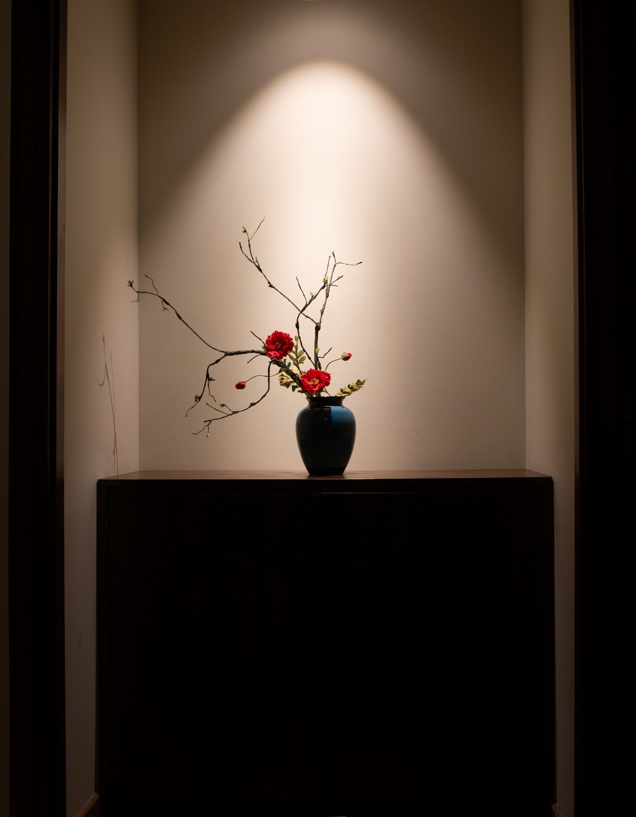 A dark wooden table with a vase containing red flowers and branches against a plain wall.